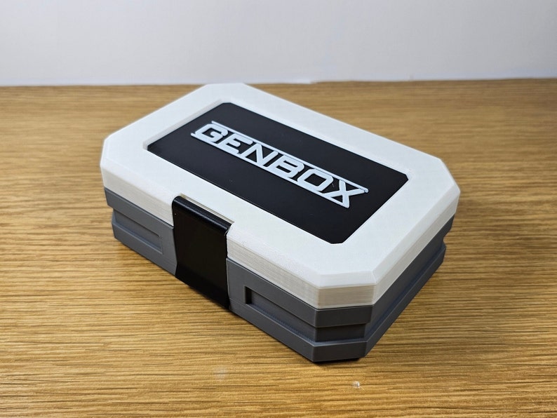 K&ouml;nnte beinhalten: Eine wei&szlig;e und graue Aufbewahrungsbox mit einem schwarzen Deckel. Der Deckel tr&auml;gt den Schriftzug "GENBOX" in wei&szlig;en Buchstaben.