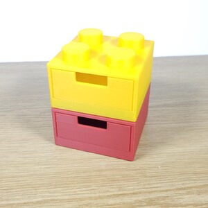 May include: Two stacked drawer organizers made to look like Lego bricks. The top drawer is yellow and the bottom drawer is red.