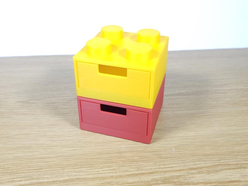 May include: Two stacked drawer organizers made to look like Lego bricks. The top drawer is yellow and the bottom drawer is red.