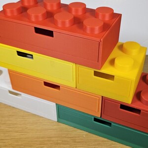 May include: A set of six colorful drawer organizers shaped like Lego bricks. The drawers are stacked on top of each other, with the top drawer being red, the second yellow, the third orange, the fourth white, the fifth green, and the bottom drawer red.