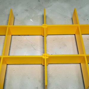 May include: A close-up of a bright yellow plastic grid structure. The grid is formed by intersecting rectangular bars, joined with interlocking components. The design creates a robust framework. The image focuses on the grid's construction and colour.