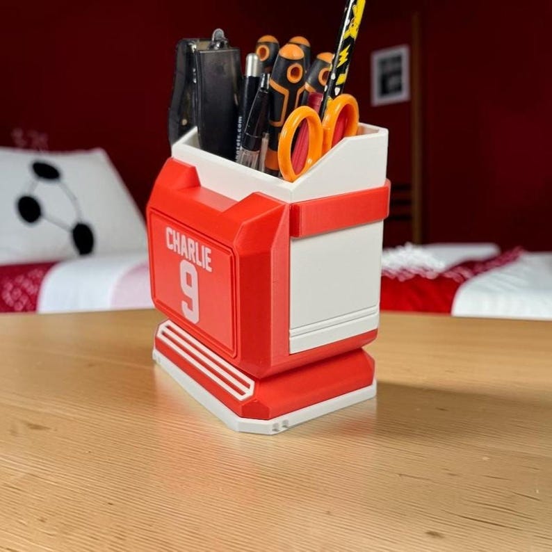 May include: A red and white desk organizer shaped like a sports jersey, featuring the name CHARLIE and the number 9 printed on the front. The container holds various office supplies, including pens, a black stapler, and orange-handled scissors.