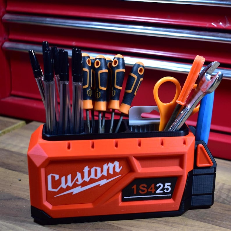 May include: An orange and black tool organizer with the word "Custom" and the numbers "1S425" printed on the front. The organizer holds pens, screwdrivers, scissors, and a wrench. The organizer is sitting on a wooden surface.
