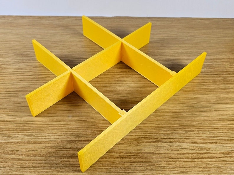 Op de afbeelding: Een gele plastic organizer met vier vakken. De organizer is kruisvormig en heeft een vierkante opening in het midden.
