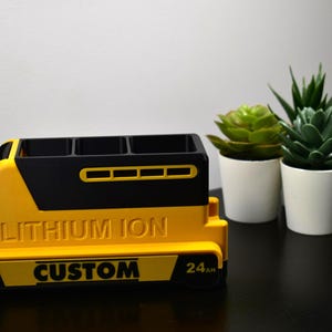Puede incluir: Un organizador amarillo y negro hecho a medida con el texto "LITHIUM ION" y "CUSTOM". El organizador tiene tres compartimentos y est&aacute; dise&ntilde;ado para parecerse a una bater&iacute;a. Dos peque&ntilde;as plantas en macetas est&aacute;n en el fondo.