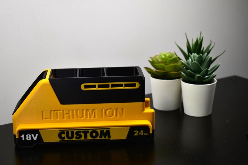 Puede incluir: Un organizador amarillo y negro hecho a medida con el texto "LITHIUM ION" y "CUSTOM". El organizador tiene tres compartimentos y est&aacute; dise&ntilde;ado para parecerse a una bater&iacute;a. Dos peque&ntilde;as plantas en macetas est&aacute;n en el fondo.