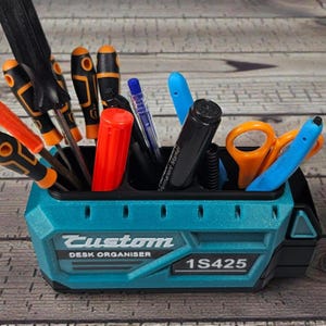 May include: A teal desk organizer shaped like a battery, filled with tools, pens, and scissors. The organizer has the text "Custom DESK ORGANISER" and "1S425" on the front. Screwdrivers with orange and black handles are visible.