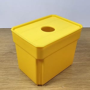 Pode incluir: Um recipiente de pl&aacute;stico amarelo com uma tampa e um furo no topo. O recipiente &eacute; projetado para conter e dispensar itens como len&ccedil;os de papel ou guardanapos.