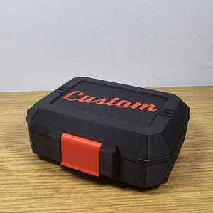 May include: A black, rectangular storage box with the word "Custom" in red script on the lid. The box has an orange clasp and is sitting on a wooden surface.