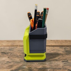 May include: A two-tone desk organiser in grey and neon green, holding pencils, scissors, and other stationery items. The organiser has a rectangular shape with a black band around the middle. The items inside are visible.