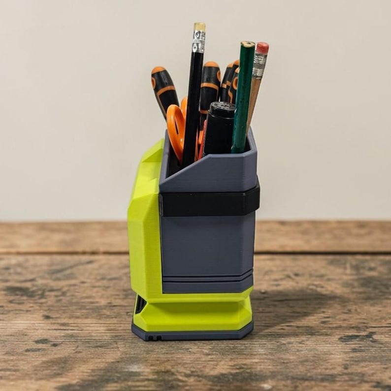 May include: A two-tone desk organiser in grey and neon green, holding pencils, scissors, and other stationery items. The organiser has a rectangular shape with a black band around the middle. The items inside are visible.