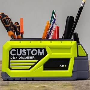 May include: A lime green and gray desk organizer with the text "CUSTOM DESK ORGANISER" in black. The organizer holds various tools and writing utensils, including screwdrivers, pens, and pencils. The product code "1S425" is also visible.