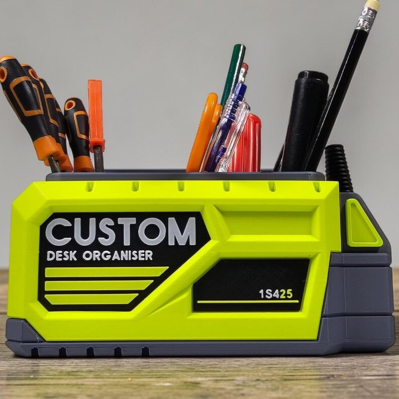 May include: A lime green and gray desk organizer with the text "CUSTOM DESK ORGANISER" in black. The organizer holds various tools and writing utensils, including screwdrivers, pens, and pencils. The product code "1S425" is also visible.