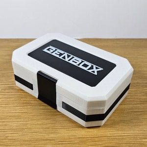 Puede incluir: Una caja rectangular blanca con una tapa negra y la palabra "GENBOX" impresa en blanco en la tapa.