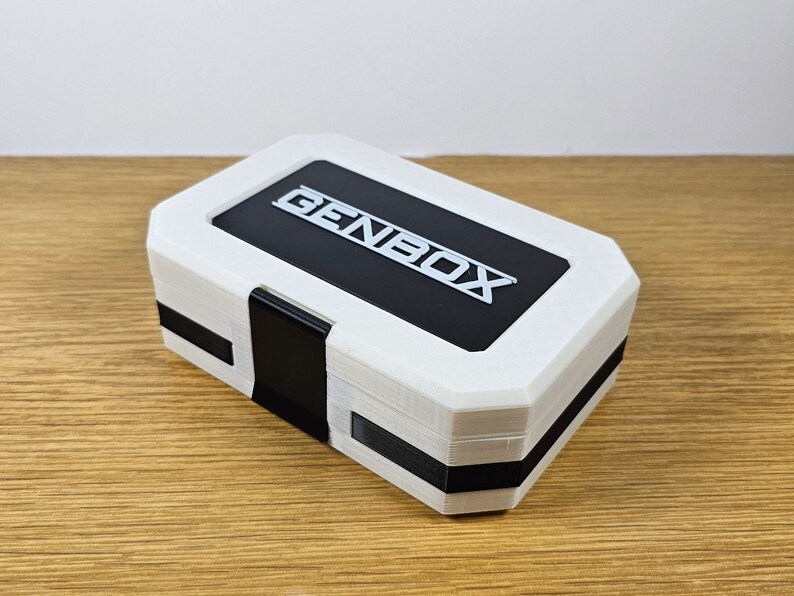 Puede incluir: Una caja rectangular blanca con una tapa negra y la palabra "GENBOX" impresa en blanco en la tapa.
