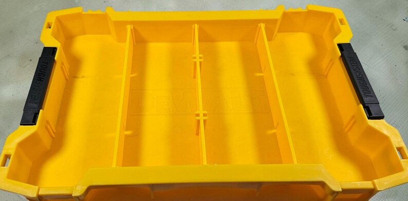 May include: Bright yellow plastic storage container with four internal dividers. The container has black handles on each end with the text "TOUGH SYSTEM". The container is designed for organising and storing tools or other items.