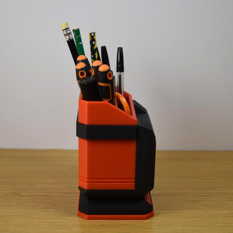 May include: An orange and black desk organizer filled with pens, pencils, and scissors. The organizer has a rectangular shape with a black band around the middle and a black base. The items inside include a pencil, pens, and orange scissors.