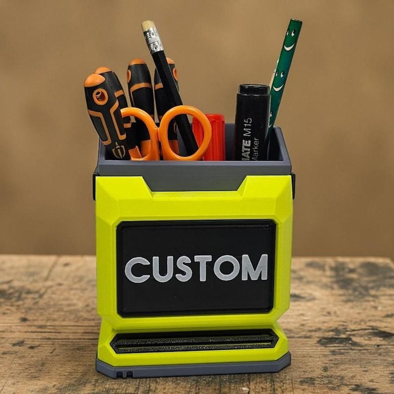 May include: A custom desk tidy in neon yellow and grey, with the word "CUSTOM" in white. It contains tools such as scissors, screwdrivers, a pencil, and a marker. The design is geometric and modern, suitable for a desk.