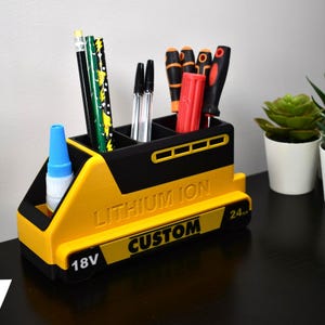 Puede incluir: Un organizador de escritorio amarillo y negro con forma de bater&iacute;a, con el texto "LITHIUM ION CUSTOM" y "18V" visibles. Contiene bol&iacute;grafos, l&aacute;pices, destornilladores y un frasco de pegamento. El organizador est&aacute; sobre una superficie oscura.