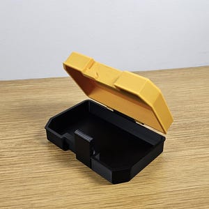 May include: A small, yellow plastic storage box with a black lid. The lid is open and the box is empty.