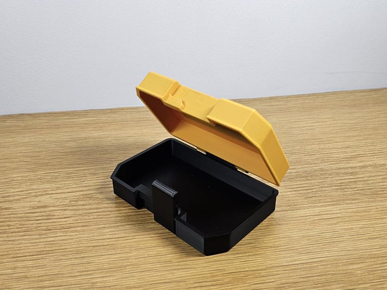 May include: A small, yellow plastic storage box with a black lid. The lid is open and the box is empty.