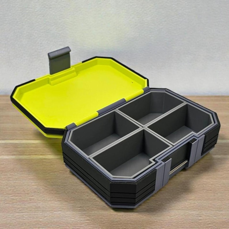 May include: A multi-compartment storage box with a hinged lid. The box is primarily gray and black, with a bright yellow interior and lid. The lid is open, revealing four interior compartments. The box is on a wooden surface.