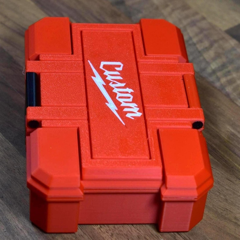 May include: A red, rectangular tool case with the word "Custom" and a white lightning bolt logo on the lid. The case has a textured surface and a secure latch. The case is sitting on a wooden surface.