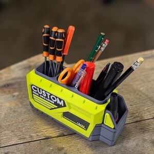 May include: A lime green and gray desk organizer with the text "CUSTOM DESK ORGANISER". The organizer holds various tools, including screwdrivers, scissors, pens, and pencils. The organizer is made of plastic and sits on a wooden surface.
