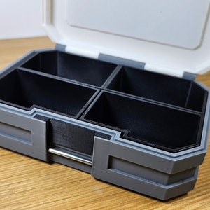 K&ouml;nnte beinhalten: Eine graue Plastik-Aufbewahrungsbox mit einem Scharnierdeckel und mehreren F&auml;chern. Die Box ist ge&ouml;ffnet und die F&auml;cher sind leer.