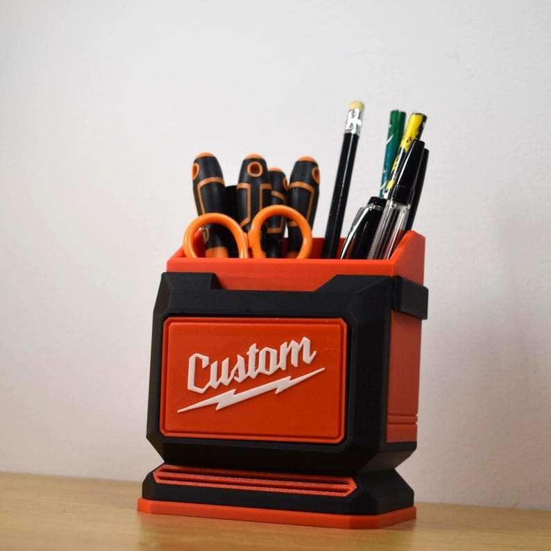May include: A red and black desk organizer with the word "Custom" in white. The organizer holds various tools, including screwdrivers, scissors, pens, and pencils. The design is a combination of red and black.