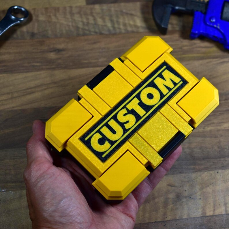以下が含まれることがあります： 黒い文字で「CUSTOM」と書かれた、黄色と黒の長方形のツールボックス。 ツールボックスは人の手で持たれています。 他のツールが背景に見えます。
