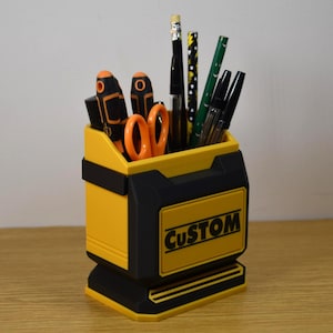 May include: A yellow and black desk organizer with the word "CUSTOM" on the front. The organizer holds various office supplies, including scissors, pens, pencils, and screwdrivers. The organizer is made of plastic and sits on a wooden surface.