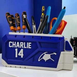 May include: A blue and white desk organizer shaped like a truck, featuring the name CHARLIE and the number 14 in white text. The organizer holds various pens, screwdrivers, and orange scissors, with a white graphic of a steering wheel on the side.