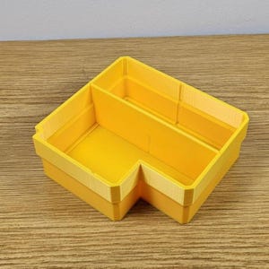 May include: A bright yellow, L-shaped desk organizer with a light yellow trim. The organizer has two main compartments and is made of a solid material. The item is sitting on a wooden surface.