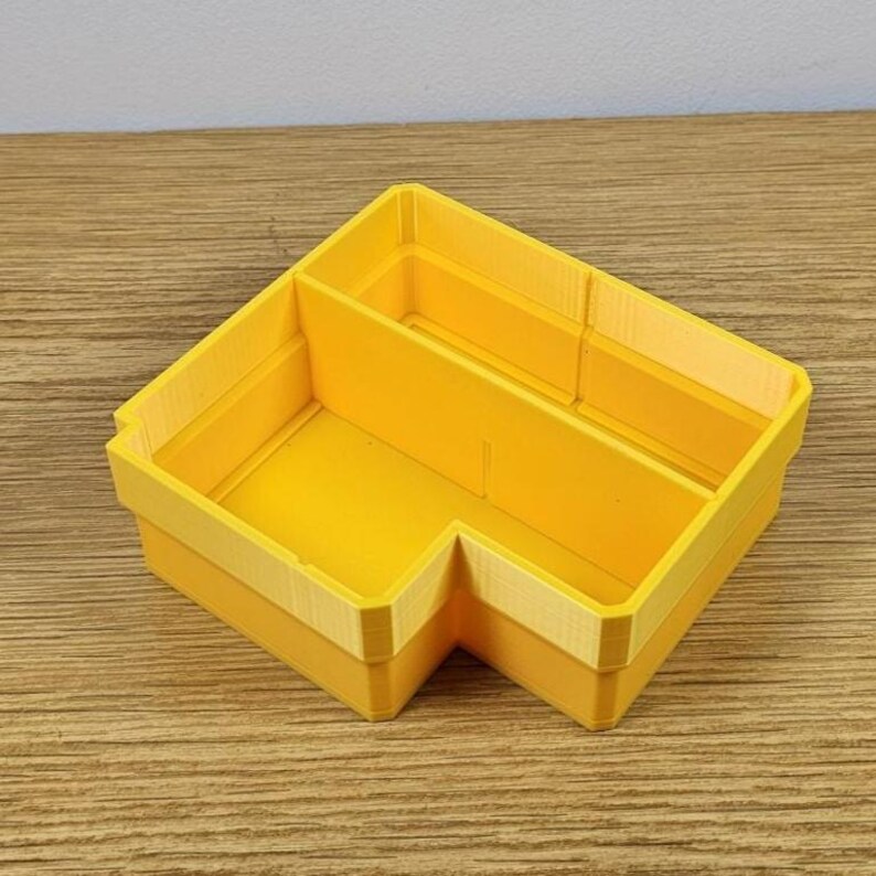 May include: A bright yellow, L-shaped desk organizer with a light yellow trim. The organizer has two main compartments and is made of a solid material. The item is sitting on a wooden surface.