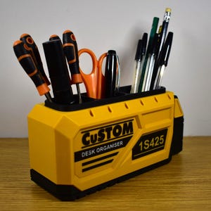 Könnte beinhalten: Ein gelber Schreibtisch-Organizer mit schwarzen Akzenten, der verschiedene Werkzeuge und Schreibutensilien enthält. Auf der Vorderseite des Organizers stehen die Wörter "CUSTOM" und "DESK ORGANISER" sowie die Nummer "1S425".