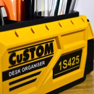 K&ouml;nnte beinhalten: Ein gelb-schwarzer Schreibtisch-Organizer mit den W&ouml;rtern "CUSTOM" und "DESK ORGANISER" auf der Vorderseite. Der Organizer hat mehrere F&auml;cher und enth&auml;lt verschiedene Werkzeuge und Schreibutensilien.