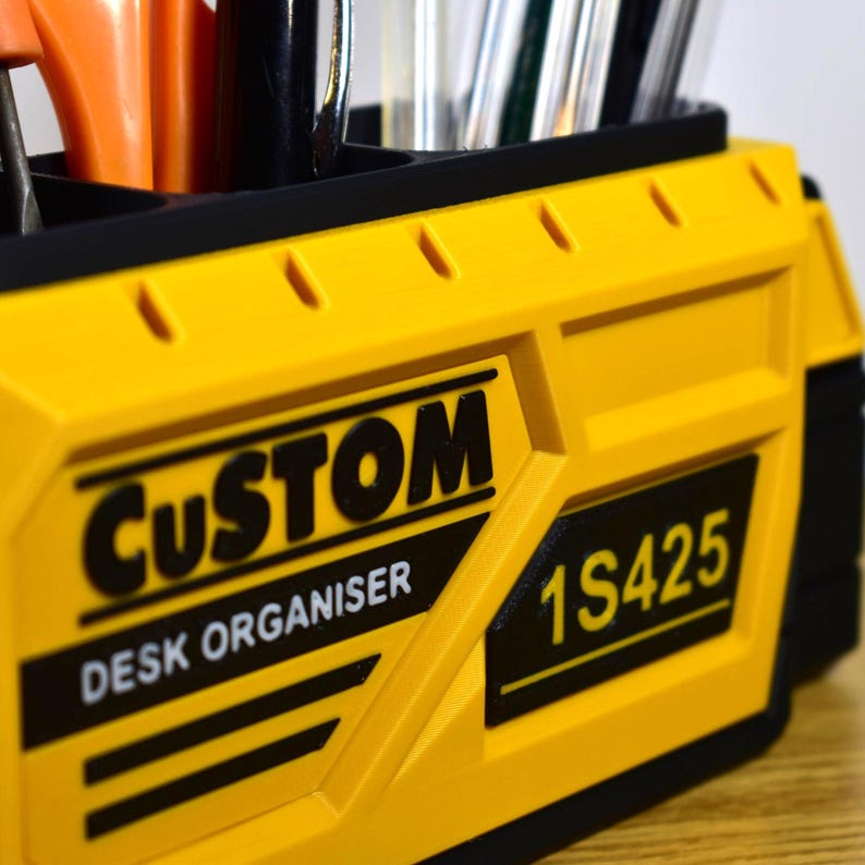 K&ouml;nnte beinhalten: Ein gelb-schwarzer Schreibtisch-Organizer mit den W&ouml;rtern "CUSTOM" und "DESK ORGANISER" auf der Vorderseite. Der Organizer hat mehrere F&auml;cher und enth&auml;lt verschiedene Werkzeuge und Schreibutensilien.