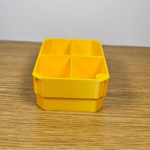 May include: A bright yellow plastic organizer with four compartments. The organizer is made of 3D printed plastic and has a smooth, slightly textured surface.