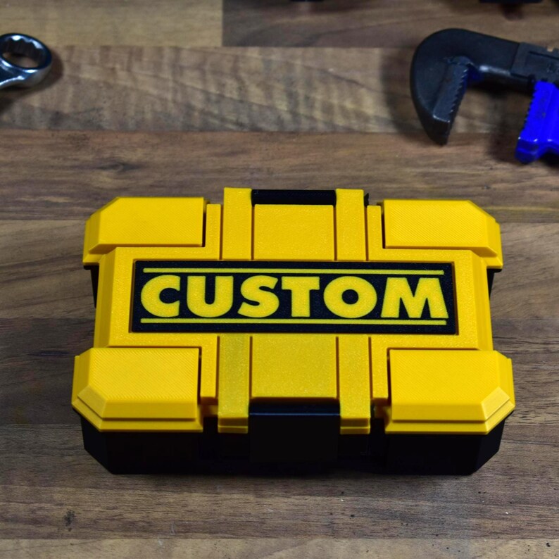 以下が含まれることがあります： 太字の黒い文字で「CUSTOM」という単語が書かれた、黄色と黒のツールボックス。ツールボックスは長方形で、ヒンジ付きの蓋と黒いベースが付いています。背景には他のツールが見えます。