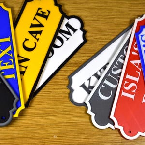 Puede incluir: Una colecci&oacute;n de letreros decorativos para habitaciones en varios colores, incluyendo negro, azul, amarillo, blanco y rojo. Cada letrero presenta un texto &uacute;nico, como "CHARLIE'S ROOM" y "MAN CAVE", en colores contrastantes.