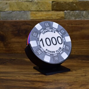 Może przedstawiać: Żeton do pokera z napisem "Charlie's 1000 Poker Club" w czarnym stojaku. Żeton jest gł&oacute;wnie szary z białymi i czarnymi akcentami, a liczba "1000" jest wyraźnie widoczna. Stojak jest czarny.