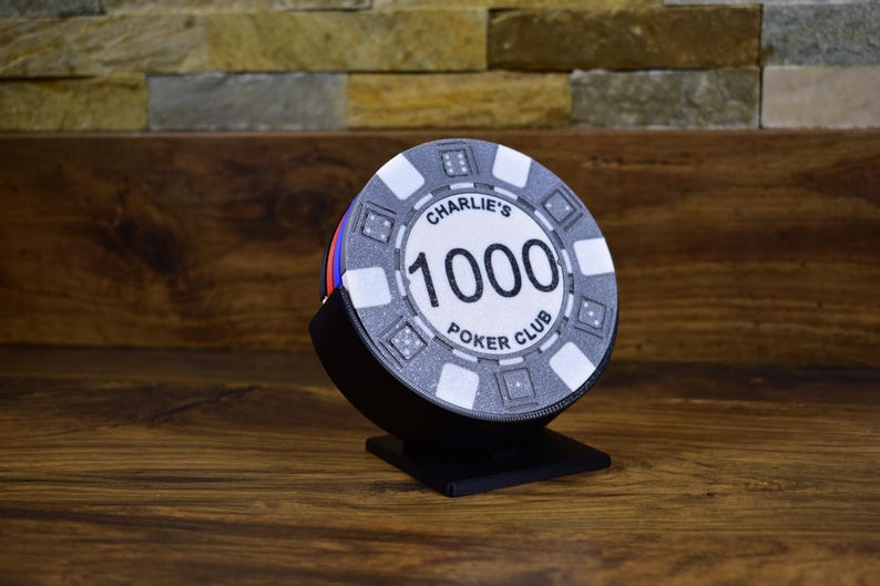 Może przedstawiać: Żeton do pokera z napisem "Charlie's 1000 Poker Club" w czarnym stojaku. Żeton jest gł&oacute;wnie szary z białymi i czarnymi akcentami, a liczba "1000" jest wyraźnie widoczna. Stojak jest czarny.