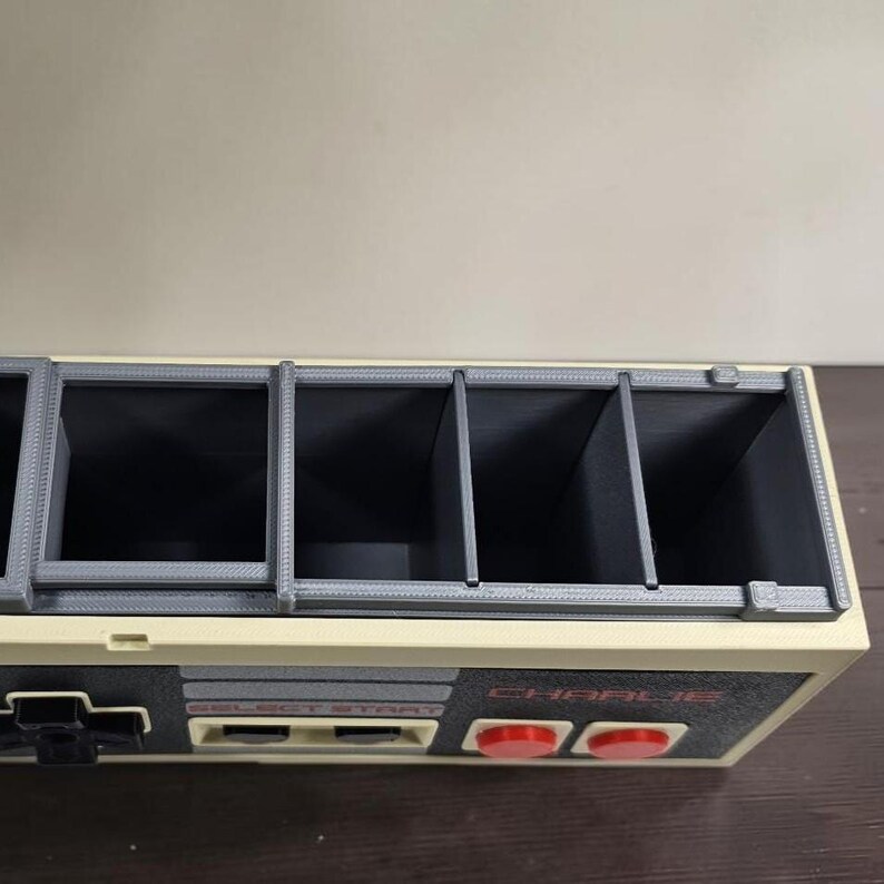 May include: A beige and gray organizer with a design inspired by a classic video game controller. It features multiple compartments and red buttons. The text "CHARLIE" is visible on the front.