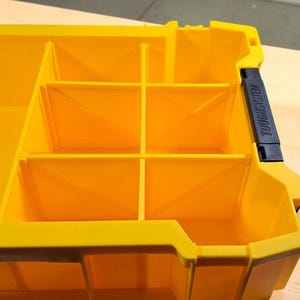 May include: A bright yellow plastic storage organizer tray featuring multiple internal compartments with diagonal structural reinforcement ribs. A black latch on the side is embossed with the text TOUGHSYSTEM in capital letters.