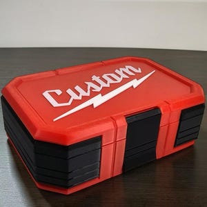 May include: A red and black rectangular storage box. The lid features the white word "Custom" in script with a white lightning bolt. The box has a textured surface and is likely made of plastic.