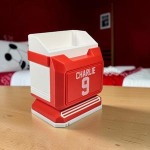 May include: A white and red desk organizer shaped like a sports jersey. The front features a raised red rectangular panel with the white text CHARLIE and the number 9 printed in white. The structure has a wide base and an open top compartment.