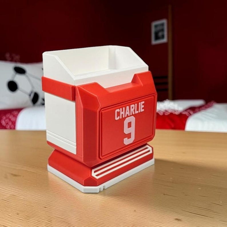 May include: A white and red desk organizer shaped like a sports jersey. The front features a raised red rectangular panel with the white text CHARLIE and the number 9 printed in white. The structure has a wide base and an open top compartment.
