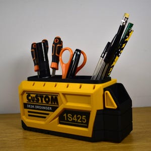 K&ouml;nnte beinhalten: Ein gelb-schwarzer Schreibtisch-Organizer mit dem Text "CUSTOM DESK ORGANISER" und "1S425". Der Organizer enth&auml;lt Schraubendreher, Scheren, Stifte und Bleistifte. Der Organizer besteht aus Kunststoff und steht auf einer Holzoberfl&auml;che.