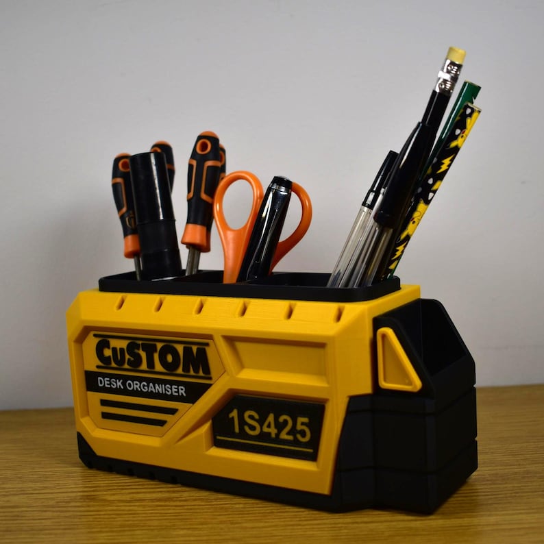 K&ouml;nnte beinhalten: Ein gelb-schwarzer Schreibtisch-Organizer mit dem Text "CUSTOM DESK ORGANISER" und "1S425". Der Organizer enth&auml;lt Schraubendreher, Scheren, Stifte und Bleistifte. Der Organizer besteht aus Kunststoff und steht auf einer Holzoberfl&auml;che.